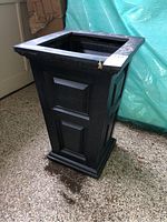 Black plastic square planter showing front side paneling and some dirt on top edge.