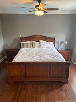 Front view of wooden sleigh bed against a gray wall showing headboard with curved top and detailed wood paneling, bed dressed with white sheets and pillows.