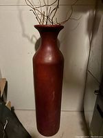 Photo of tall solid red vase with narrow neck and flared mouth, with some dried twigs inside, standing against plain wall.