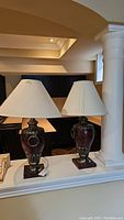 Pair of ceramic base table lamps with fabric shades shown together on ledge. Both lamps have matching dark burgundy ceramic bases with metallic detailing and off-white fabric shades.