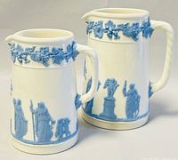 Two Wedgwood creamers side by side showing blue relief classical figure design, handled and spouted, cream base color.