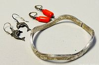 Photo showing silver bangle bracelet with wave design and two pairs of earrings, one orange-red bead pair and one silver dolphin pair.