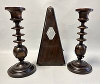 Front view of the vintage wooden metronome flanked by two turned wooden candleholders on each side, on a plain background.