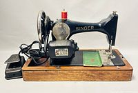 Full side view of black Singer 128-13 electric sewing machine on wooden base with foot pedal and green instruction manual beside it.