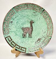 Front view of green patina copper plate with silver inlay camel and rim design on wooden stand.