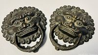Pair of cast metal door knockers with stylized lion face and ring handles shown side by side.