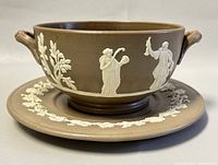 Side view of the Wedgwood jasperware brown double-handle soup bowl on matching saucer showing white classical figures in relief on bowl and grapevine motif on saucer.