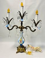 Full view of vintage 3-arm Italian porcelain and brass electric candelabra with floral hand-painted details and hanging crystal drops, amber flame bulbs, and coiled electric cord.