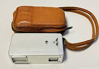Side view of the silver vintage Minolta-16 camera with brown leather case beside it, showing the camera's rectangular metal body and part of the leather case with wrist strap.