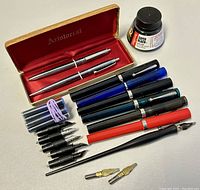 Photo showing assorted vintage Sheaffer fountain pens in black, blue, teal, red, with nibs laid out, ink bottle labeled Super-Black Ink, bundles of refill cartridges, extra nibs, and a dip pen holder.