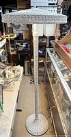 Full view of vintage white wicker floor lamp with a round woven lampshade and base, standing on the floor among other items.