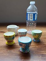 Front view showing all five ceramic egg cups with colorful bird and animal decorations and one plain white with legs, alongside water bottle for scale.