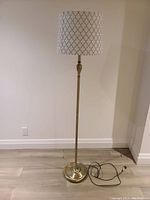 Full view of tall floor lamp with light brass colored metal base and stand, and white fabric lampshade with black geometric pattern.