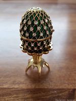 Front view of the green enamel egg-shaped trinket box with gold lattice and clear rhinestones on a gold claw base stand.