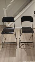 Two Ikea folding barstools from front view, showing black-brown wood veneer seat and backrest, and powder coated steel legs.