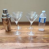 Photo of four clear martini glasses arranged in two rows alongside a stainless steel shaker and a water bottle for size reference on a wooden surface.