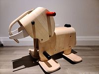 Left side angle view of the wooden elephant ride-on toy showing smooth light wood finish, red handlebar, black footrests, and white tusks with a minimalist design.