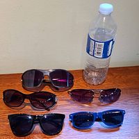 Five pairs of sunglasses displayed on a wooden surface next to a water bottle for size reference.