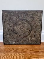 Front view of square wood carved wall art on hardwood floor showing geometric circular pattern.