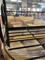 Close-up of the side and shelf bars of the black metal baker's rack wrapped partially in plastic wrap, showing shelf rods and decorative swirl design.