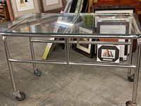 Overall view of rectangular cart with clear glass top, metal tubular frame, and four wheels.