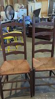 Two antique wooden ladder back chairs with woven rush seats shown together in storage setting.