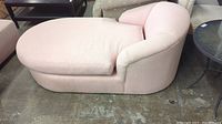 Photo of chaise lounge showing full body from side angle, light pink upholstery, rounded edges, fabric seat cushion, placed on concrete floor.
