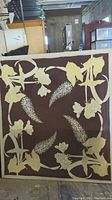 Full view of the fabric botanical wall art showing cream floral and leaf designs on a brown background, with visible hole and damage.