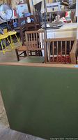 Photo showing the four wooden chairs arranged behind the folding green felt game table with wooden framing, in a cluttered storage area.