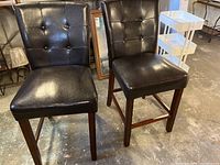 Two black leatherette bar stools with padded seats and backs, standing on a concrete floor in a warehouse. Wooden legs with footrests. Upholstery shows visible signs of wear.