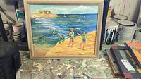 Full view of framed mixed media beach scene painting showing water, rocky shore, and several figures near the water; piece is resting on a paint-splattered surface with paint cans in the background.