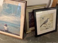 Three framed printed pictures leaning against a wall on warehouse floor showing different scenes: a boy by the sea, a bird and butterfly near a birdcage, and an ancient monks image.