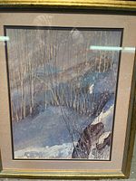 Full view of one framed snowy nature scene artwork showing bare trees and snow with gold frame and beige mat.
