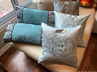 Five cushions arranged on a chair showing two rectangular teal and patterned cushions plus three square embroidered floral cushions.