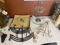 Decorative square plate with bird design and set of black curved metal plate stands and gold metal plate hangers on table; two-tier glass serving dish next to them.