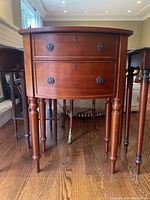 Front view of the wooden end table with two drawers and four turned legs on a wooden floor.