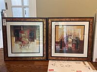 Two framed abstract prints side by side on a wooden surface. Each print is 29x29 inches with warm color abstract designs.