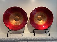 Pair of red and gold tinted Murano glass bowls displayed on black metal stands, showing reflective and textured design.