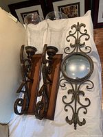 Photo of two metal and wooden wall sconces with clear hurricane glass candle holders, and one ornate round metal-framed mirror lying on a white surface.