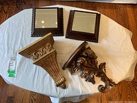 Two square wooden framed mirrors and two painted wood decorative wall sconces on white sheet, displayed on wooden floor.