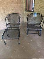 Two black metal outdoor armchairs with one matching ottoman placed on a concrete patio floor against a stucco wall.
