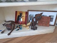 Two metal cat statues with green eyes, all cat decorations arranged on a shelf with framed pictures behind.