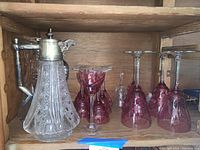 Two crystal pitchers displayed alongside rose colored wine and sherry glasses in wooden cabinet.