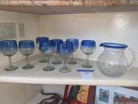 Pitcher and eight matching glasses lined up on shelf, clear glass with blue accents