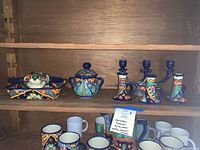 Top shelf showing a teapot with matching mugs, ceramic serving dish with cover, and three Talavera candlesticks (one three-armed and two single.) Below are additional mugs in various colors and designs.