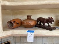 Three wooden decor pieces on a shelf: a hand-painted vase with braided rim on left, a round wooden vase with geometric circles in center, and a dark wooden tiger sculpture on right.