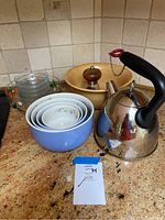 Photo showing tea kettle, set of nested mixing bowls, wooden salt/pepper mill on wooden bowl base, and six glass cups.