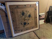 Framed painted cloth art featuring a bird and blue floral design on beige cloth, square shape.