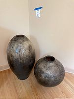 Two large ceramic vessels placed on a wooden floor against a neutral wall, showing the size difference and finish.