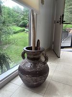 Large brown jug style vase with horizontal white stripes and decorative tile inserts, containing two natural decor sticks, placed on tiled floor near window with view to green outdoor path.